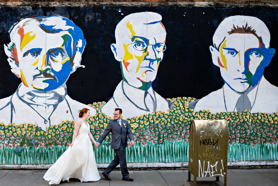 A newly married couple walk along the street holding hands in front of a colorful mural in Ann Arbor, MI.